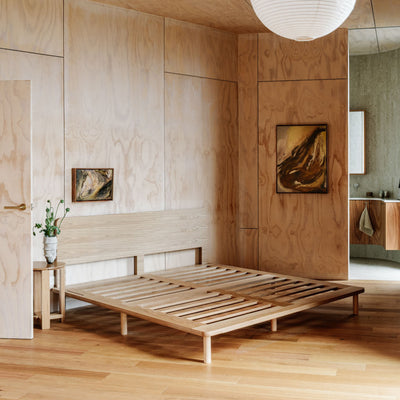 A minimalist light oak wooden bed frame with a slatted platform base and matching headboard, styled in a modern, warm-toned bedroom with plywood walls and wood flooring.